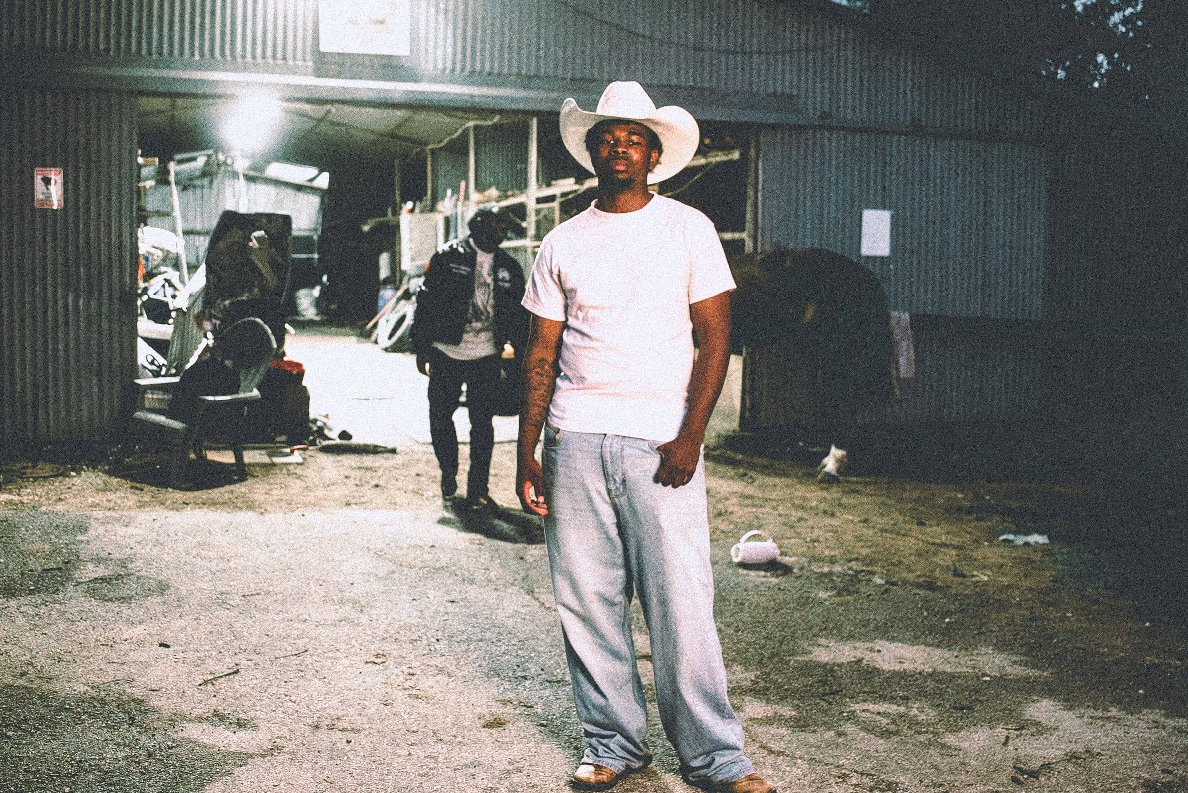 Luhdeeskii in a white cowboy hat standing outside a tin barn at night, lit by a single work light